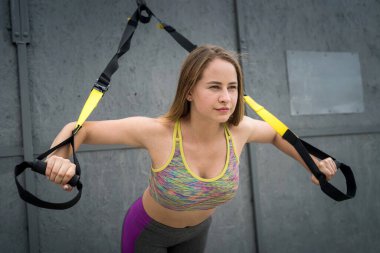 Spor kıyafetli genç bir kadın doğada trx fitness kayışlarıyla şınav çekiyor. Sağlıklı yaşam tarzı