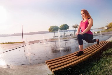 Güzel genç bir kadın sabah parkta çalışmadan önce esneme egzersizi yapıyor.