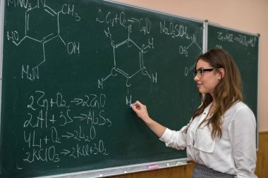 Gözlüklü komik bayan öğretmen bilimsel formüllü tebeşirle tahtaya karşı. kimya