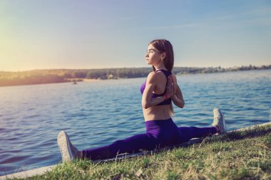 Genç Avrupalı bayan sporcu bacaklarını esnetme egzersizleri yapıyor, dışarıda göl kenarındaki yarıklara oturuyor. Sağlıklı yaşam tarzı