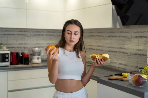 Sporty woman thinking what eat on breakfast between fruits and sweet bun on her kitchen. Concept of healthy and unhealthy food