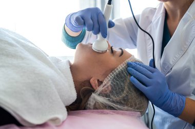 Klinikte havlunun altında kanepede yatan genç bir bayan için fonforez prosedürü uyguluyor. Kozmetoloji ve profesyonel cilt bakımı.