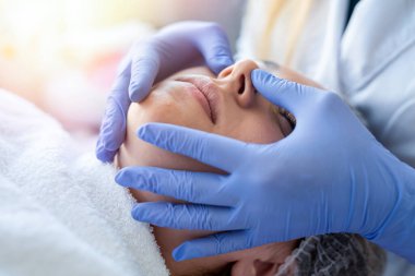 Kozmetik uzmanı mavi eldivenleri verdi. Masaj yapan genç bir hastanın yüzüne dokundu. Cilt bakımı ve gençleştirme kavramı.