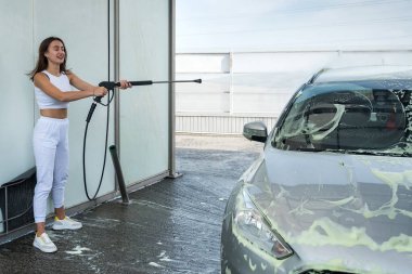 Güzel bir kız oto yıkama istasyonunda arabasından çıkan su tabancasıyla köpüğü yıkar.