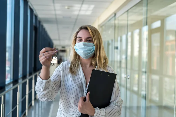 Smiling woman with protective mask with house keys in modern business center. Concept of real estate and office