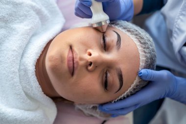 Klinikte havlunun altında kanepede yatan genç bir bayan için fonforez prosedürü uyguluyor. Kozmetoloji ve profesyonel cilt bakımı.