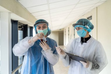 Tıbbi koruyucu giysiler giyen çift hemşire modern klinikteki test tüpünde kan örnekleri tutuyor.
