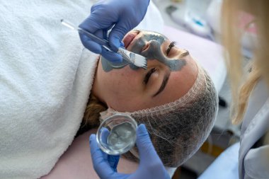 Kozmetik uzmanı, kaplıca salonundaki genç ve güzel bayana siyah maske yapıyor. Öncül cilt bakımı prosedürü.