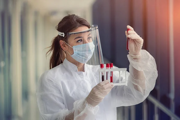 beautiful young nurse in protective mask and face shield and uniform examines blood samples in test tube for coronavirus infection. covid19