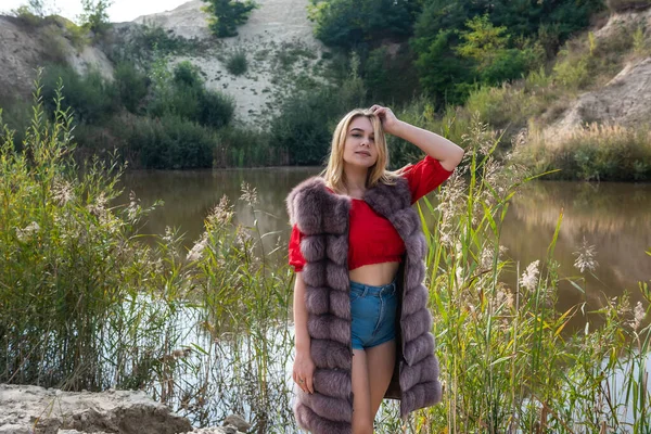 beautiful girl in a fur coat and a light red T-shirt and shorts posing by the lake on a warm autumn day