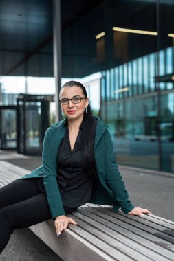 Beyaz takım elbiseli güzel bir kadın şehrin dışında oturuyor. Mavi cam binanın arka planında.