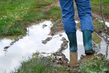 Yağmurdan sonra çamurlu bir yolda yürüyen yeşil lastik çizmeler ve kot pantolon giyen bir adam. Kırsal yaşam tarzı, çiftçilik, açık hava işi veya kırsal macera kavramı.