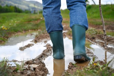 Çamurlu, su basmış bir yolda yürüyen yeşil lastik çizmeli bacaklar. Açık hava macerası ve kırsal yaşam tarzı konsepti.
