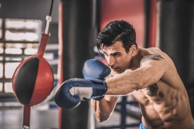 Spor salonunda egzersiz yapan kaslı bir adam kum torbasını tekmeliyor.