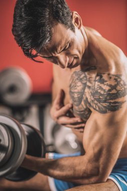 Göğsünde dövme olan genç bir adam spor salonunda halterlerle çalışıyor.