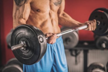 Göğsünde dövme olan genç bir adam spor salonunda halterlerle çalışıyor.