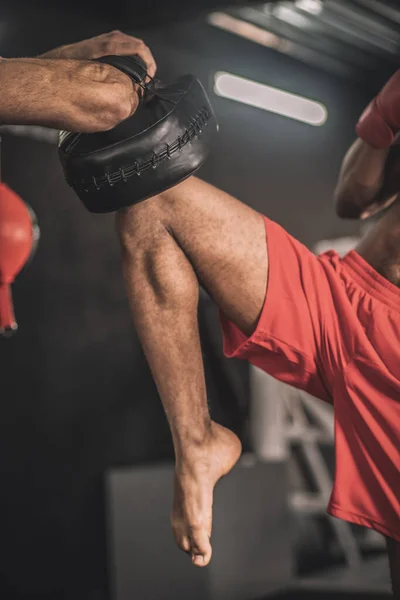 Koyu renk saçlı genç boksör bir koçla bacak tekmesi üzerinde çalışıyor. — Stok fotoğraf