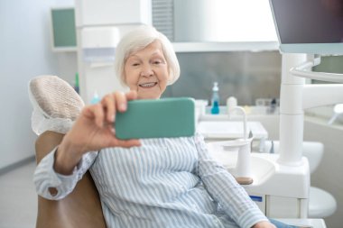 Gri saçlı kadın diş yenileme ameliyatından sonra selfie çekiyor ve memnun görünüyor.