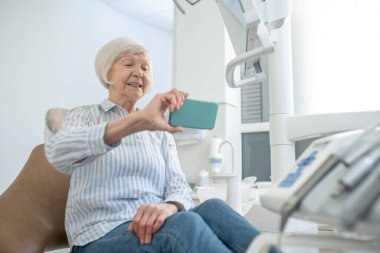 Gri saçlı kadın diş yenileme ameliyatından sonra selfie çekiyor ve memnun görünüyor.