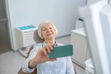 Gri saçlı kadın diş yenileme ameliyatından sonra selfie çekiyor ve memnun görünüyor.