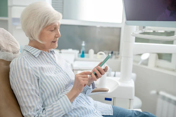 Mulher sênior sentada no consultório de dentistas e usando seu smartphone — Fotografia de Stock