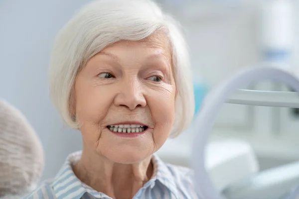 Senior woman sitting at the dentists office and looking in the mirror after teeth restoration — Φωτογραφία Αρχείου