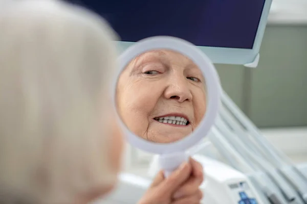 Senior mulher sentada no consultório de dentistas e olhando no espelho após a restauração dos dentes — Fotografia de Stock