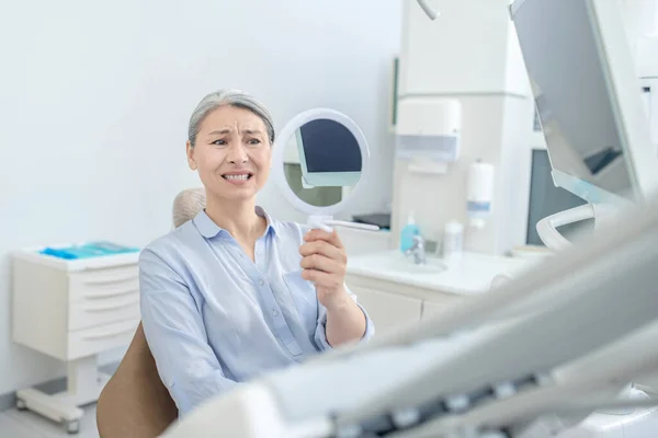 Mulher sentada no consultório de dentistas com um espelho nas mãos — Fotografia de Stock