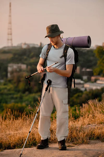 A young man with backpack standing on the hill with walking sticks in hands — Stock Photo, Image