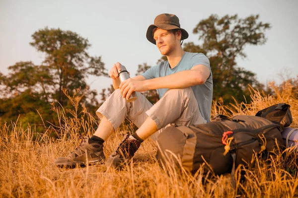 Ένας άντρας με καπέλο κάθεται στο λόφο και ανοίγει το θερμός του — Φωτογραφία Αρχείου