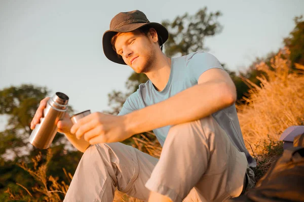 Ein Mann mit Hut sitzt auf dem Hügel und öffnet seine Thermoskanne — Stockfoto