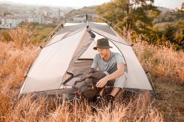 Çadırın yanında oturan şapkalı bir adam sırt çantasını topluyor.