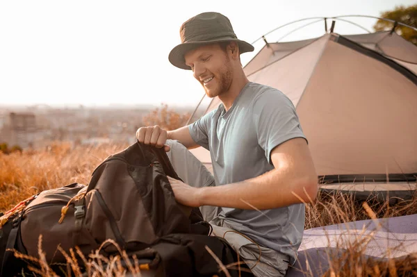 Человек в шляпе пакует рюкзак, сидя возле палатки. — стоковое фото