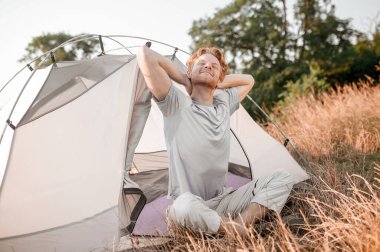 Kızıl bir adam çadırın yanında oturuyor ve uykulu ve rahat hissediyor.