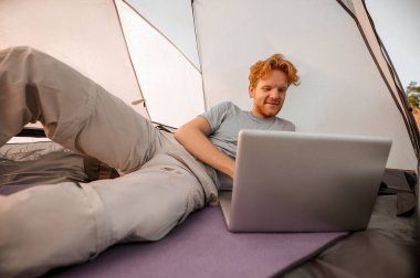 Genç bir adam elinde dizüstü bilgisayarla çadırda yatıyor.