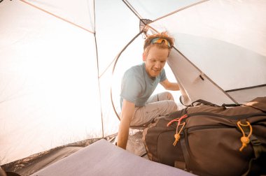 Çadırda neşeli görünen bir delikanlı sırt çantasının yanında oturuyor.