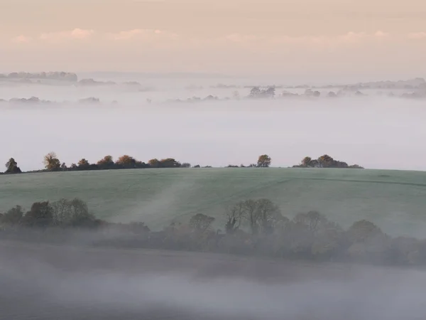 Dense fog in the dips surrounding Old Winchester Hill with farmers fields in the foreground. Colours are vey pastel like due to the fog.