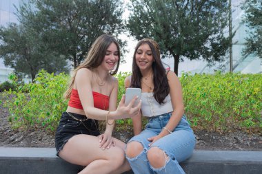 Parkta konuşan iki genç bayan bir saksı hakkında, bahar günü yeşil ağaçlar ve mavi gökyüzü, bulutsuz