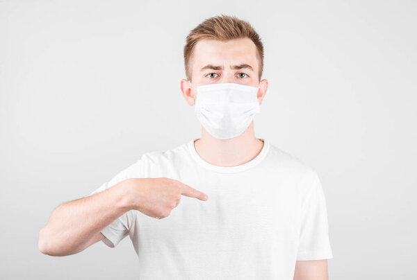 Portrait of young man in white shirt with surgical medical mask standing, poinitng himself and looking at camera.