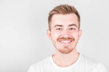 Beyaz gömlekli, kendine güvenen genç, yakışıklı bir adam beyaz arka planda dururken gülümsüyor..