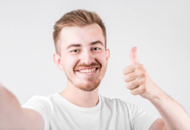 Sıradan tişörtlü yakışıklı bir adamın fotoğrafı, kameraya baş parmağıyla gülümserken beyaz arka planda izole edilmiş selfie çekiyor..