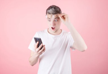 Şok olmuş beyaz tişörtlü bir gencin portresi. Pembe arka planda izole bir şekilde cep telefonuna bakıyor.