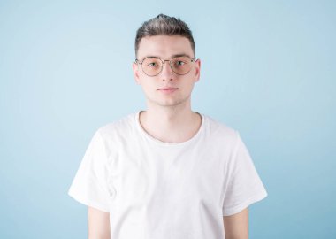 Şık bir saç stili olan, sıradan beyaz tişört giyen, stüdyoda mavi bir arka planda poz veren, münferit bir genç adamın görüntüsü ciddi bir ifadeye sahiptir.