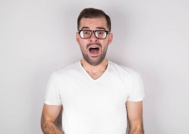 Genç adam, ağzı ve yuvarlak gözleri örtüyor, yuvarlak gözlükler takıyor, derin şaşkınlık ve korku yaşıyor, gri arka planda izole edilmiş.