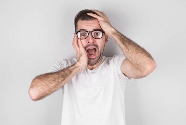 Genç adam elleri ve yuvarlak gözlükleriyle ağzını kapatıyor. Gri arka planda derin şaşkınlık ve korku yaşıyor.