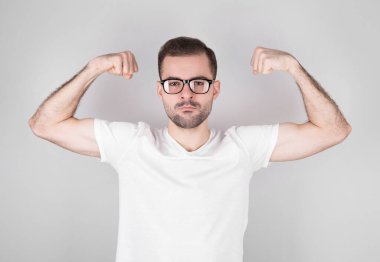 Gri arka planda tişörtü olan genç yakışıklı bir adam güçlü bir adamdır. Kollarının kaslarını gösterir. Kendinden emin ve gururludur.