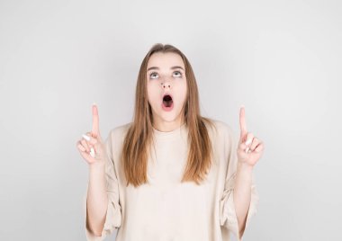 Bej tişörtlü şaşırmış bir kız yukarı bakar, gri arka planda ağzını açar.