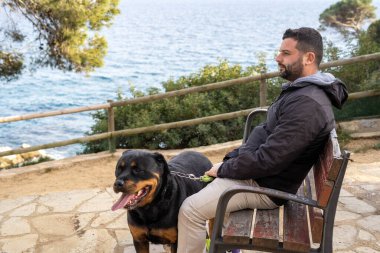 Parktaki bankta oturan adam Rottweiler köpeğinin yanında.