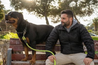 Parktaki bankta oturan adam Rottweiler köpeğinin yanında.