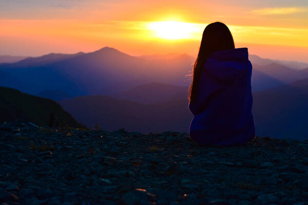 A girl in a warm hoodie sits on the edge of a cliff in the mountains at sunset